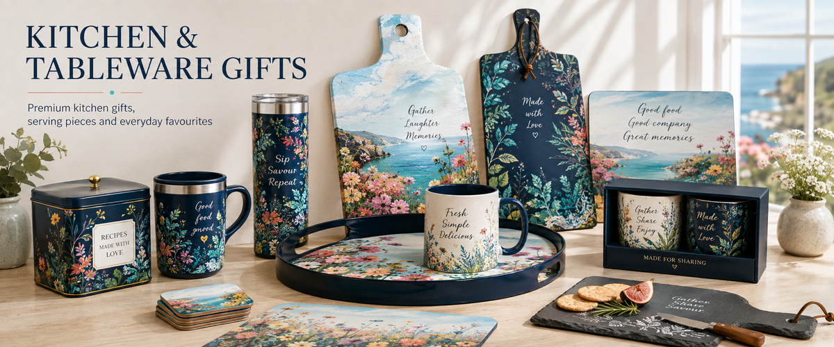 A styled kitchen and tableware gift set displayed on a tabletop with a window and flowers in the background. The set includes floral ceramic mugs, a tin canister, a travel mug, a serving tray, cutting boards, coasters, and a boxed candle set.