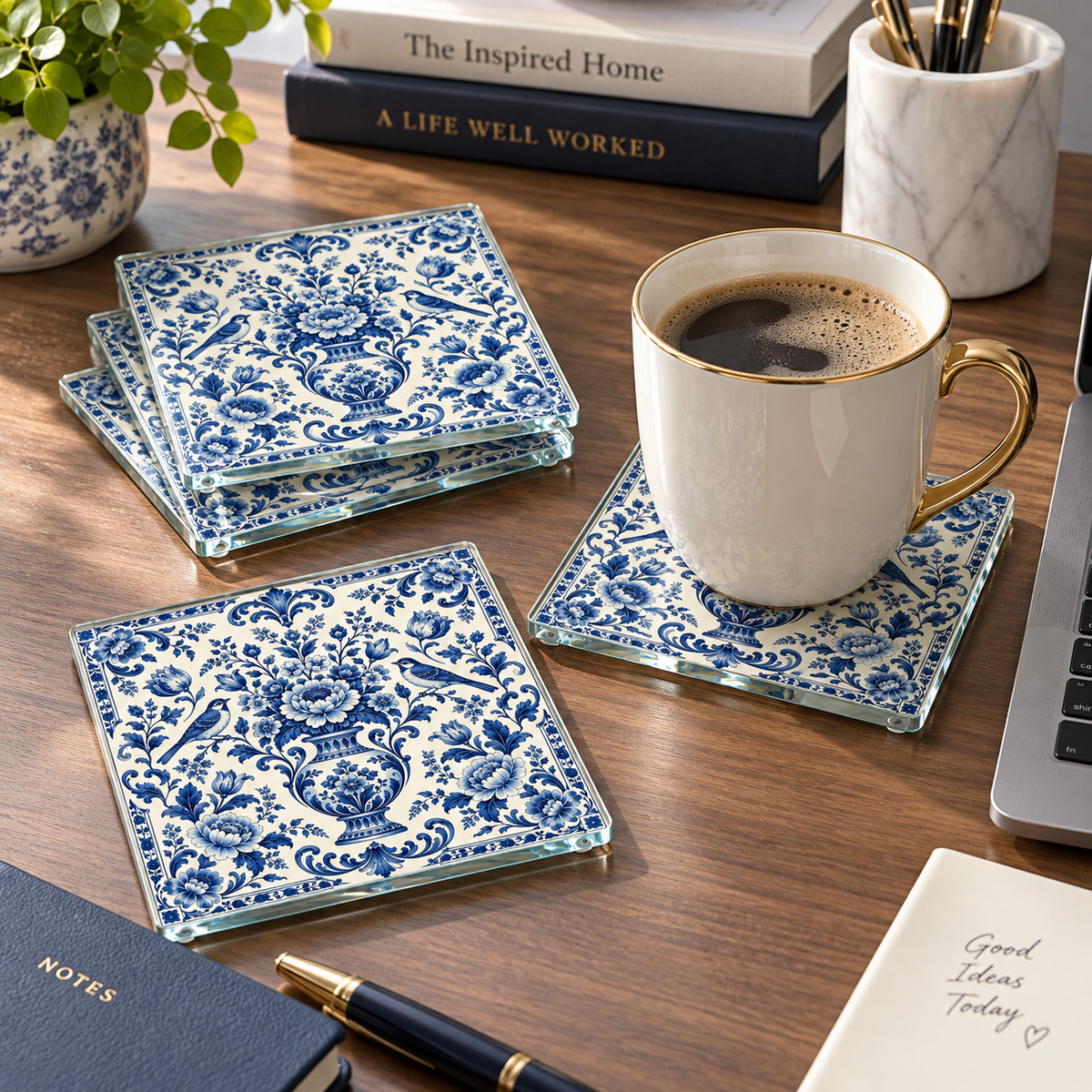 Four square glass coasters with a blue and white floral pattern arranged on a wooden desk, with a white coffee mug on one coaster. Other desk items including books, a plant, a laptop edge, and a pen are visible in the background and foreground.