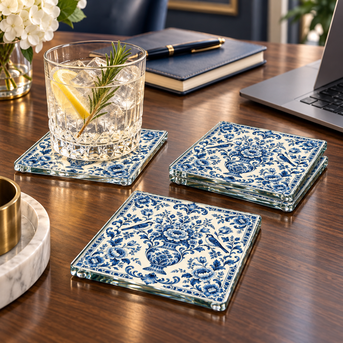 Three square glass coasters with a blue floral pattern arranged on a wooden desk, with a drinking glass and other desk items nearby.