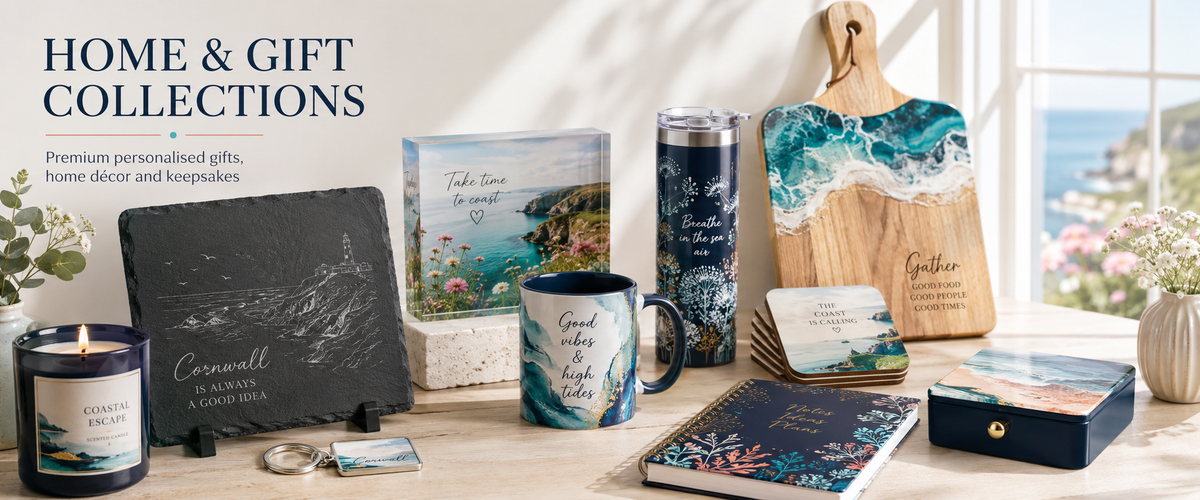 A styled collection of home and gift items displayed on a tabletop in front of a bright window. The products include a slate sign engraved with Cornwall, a navy candle jar, a ceramic mug with a blue and white abstract print, a wooden cutting board with ocean artwork, a tumbler, coasters, a notebook, a small box, and a candle.