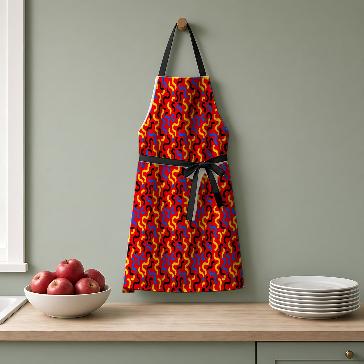 Squiggle Red Apron hanging on a sage-green kitchen wall