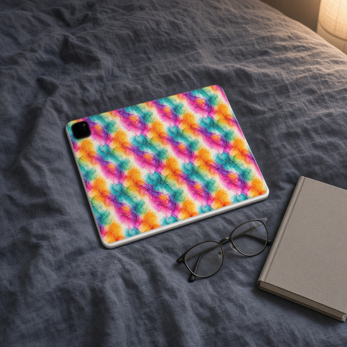 Rainbow Alcohol Ink Bloom iPad folio case on slate-blue bedding with a book and glasses