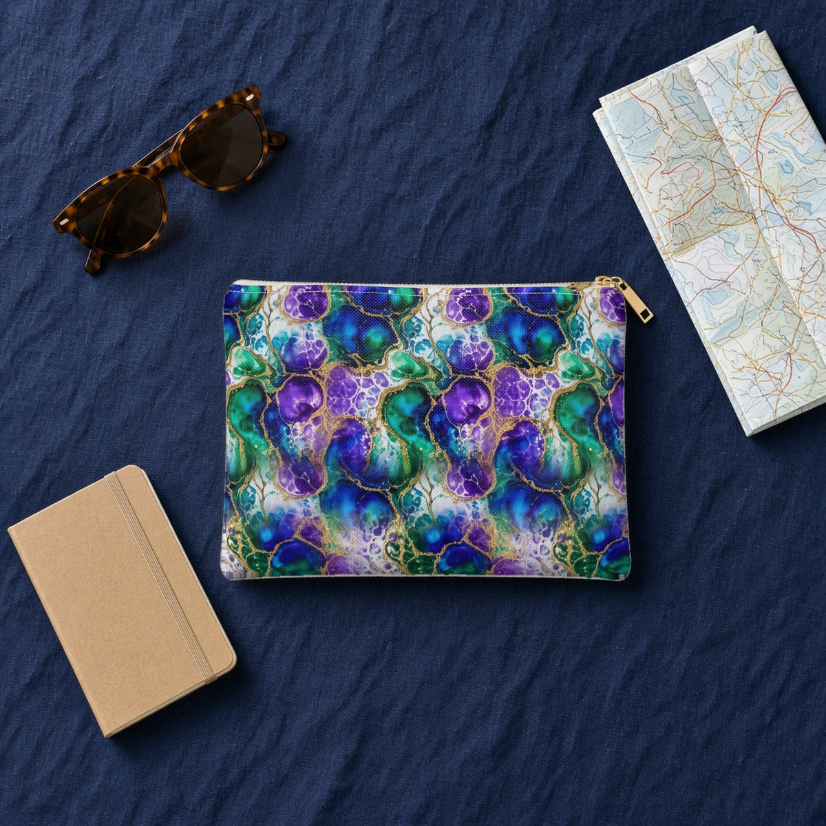 Peacock Acrylic Pour Cells zip accessory pouch on navy linen with sunglasses, a notebook and map