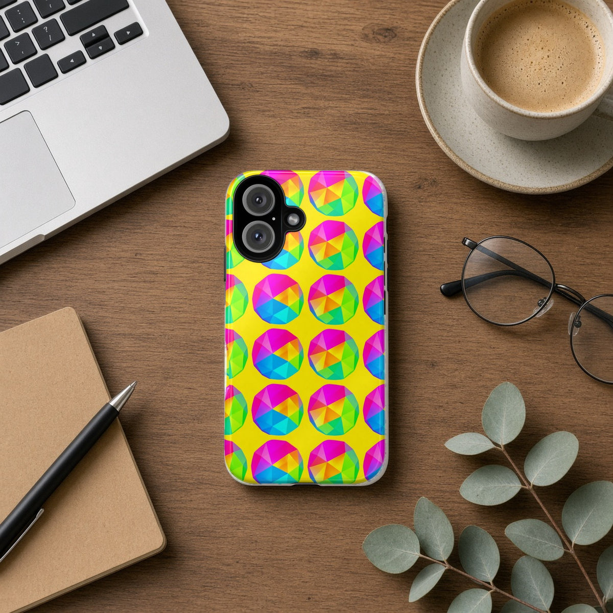 Holo Pebble Prisms tough phone case on a wooden desk beside a laptop, coffee and eucalyptus