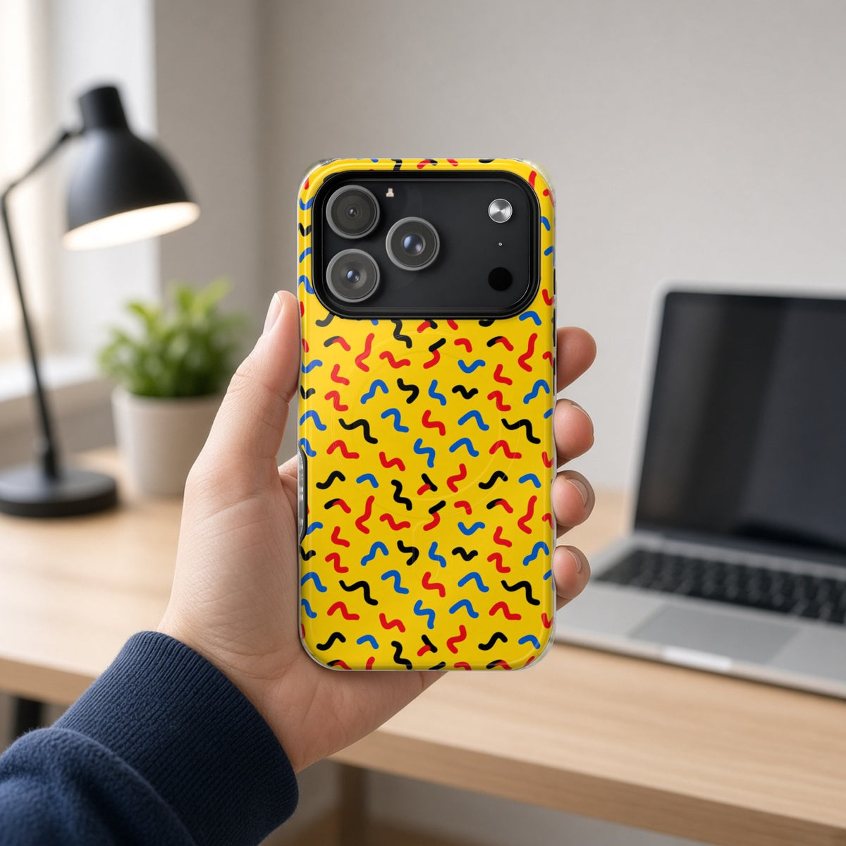 Hand holding the Squiggle Strokes Yellow magnetic iPhone case at a home office desk